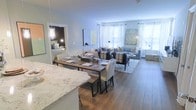 This well-appointed single-floor residence is designed around an open-concept layout that connects the kitchen, dining, and living areas in one cohesive, flowing space. The kitchen features a striking granite countertop with a light, veined finish, offering both beauty and functionality as it opens directly to the main living area. A rustic wood dining table with upholstered white chairs and a coordinating bench anchors the dining zone, providing an inviting space for gathering and meals. The living area is generously proportioned, centered around a comfortable sectional sofa and accented with layered textiles, patterned throw pillows, and a classic chevron area rug that adds visual interest underfoot. Rich hardwood flooring runs throughout, lending warmth and continuity to the space. Large windows dressed with sheer white curtains line the far wall, flooding the interior with natural light and creating an airy, welcoming ambiance. Thoughtfully placed lamps and wall art add character and a curated aesthetic throughout. A glimpse into an adjacent bedroom reveals a well-lit, comfortably furnished room that maintains the same refined and approachable design sensibility found throughout the rest of the home.