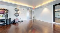 Located at 1600 Harbor Blvd in Weehawken, NJ, this fitness and dance studio offers a thoughtfully designed single-floor space ideal for a variety of movement-based activities. The studio is defined by its rich, dark hardwood flooring that spans the entire room, providing both durability and an elegant aesthetic underfoot.

A full wall of floor-to-ceiling mirrors runs along one side of the studio, complete with mounted wooden ballet barres, making the space well-suited for dance, barre, yoga, or stretching classes. The mirrors also enhance the sense of openness, visually expanding the room. A flat-screen TV is mounted within the mirrored wall section, offering instructional or entertainment capabilities during sessions.

Large, multi-pane windows line an entire wall, flooding the studio with generous natural light and offering views of the surrounding Weehawken neighborhood. The bright, airy atmosphere is further enhanced by warm cove lighting integrated into the tray ceiling, creating an inviting and motivating environment.

A frosted glass sliding door provides a stylish and functional entry point, maintaining privacy while contributing to the modern design. The studio also accommodates fitness equipment such as stability balls, reflecting its versatility for multiple workout disciplines. Clean white walls and recessed lighting complete the polished, contemporary look of this well-equipped space.