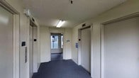 This elevator lobby serves as a central circulation hub within the building, providing access to multiple elevator bays along with a doorway leading to an exterior area. The space is finished with dark blue-grey carpeting underfoot, offering a practical and durable surface suited for high-traffic use. The walls are finished in a neutral off-white tone, contributing to a clean and functional aesthetic throughout the area. Overhead, a textured ceiling with fluorescent lighting ensures the space is well-illuminated, supporting safe and comfortable navigation. Two elevator doors are prominently positioned on either side of the lobby, each accompanied by standard call panels and informational signage. An interior door centrally located leads to an exterior-facing transitional space, allowing natural light to filter into the lobby and providing an additional point of egress. The overall layout is straightforward and efficient, prioritizing ease of movement and accessibility. Informational placards are mounted on the walls, offering building-related guidance to occupants and visitors. The space reflects a utilitarian design approach consistent with a professional or residential multi-floor building environment.