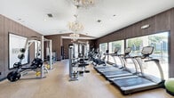 Located at 915 Lovvorn Rd in Carrollton, Georgia, this fitness center at River Pointe offers a thoughtfully designed exercise space that balances functionality with refined style. The room features vaulted ceilings adorned with a striking crystal chandelier, creating an unexpectedly elegant atmosphere for a workout facility. Warm wood-paneled walls line the interior, contributing to a sophisticated and cohesive aesthetic throughout.

The space is comprehensively equipped to support a full range of workout routines. Cardio equipment includes multiple treadmills and stationary bikes positioned along a large window that allows natural light to pour in and provides views of the outdoors. A dedicated free weights area features a full rack of dumbbells alongside adjustable benches, while multiple cable and strength machines are distributed throughout the floor. Rubber flooring tiles are strategically placed beneath equipment to provide stability and protection.

A built-in storage and service area along one wall includes overhead cabinetry, a countertop workspace, and a mini-refrigerator, offering added convenience for users. Large mirrors expand the sense of space and allow for proper form checks during exercise. The room is well-lit with recessed lighting supplementing the natural light, and sound equipment is mounted on the walls. Multiple entry and exit points ensure ease of access throughout the facility.