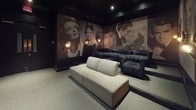 Located at 102 Fallsgrove Blvd in Rockville, Maryland, this single-floor Hollywood-themed screening lounge delivers a cinematic experience steeped in classic film nostalgia. The space is defined by dramatic floor-to-ceiling murals depicting iconic Hollywood legends — including Marilyn Monroe, James Dean, Humphrey Bogart, Audrey Hepburn, and scenes from "Gone with the Wind" — printed in rich sepia and black-and-white tones that wrap the dark walls in a gallery-like fashion.

The interior is anchored by deep charcoal and matte black surfaces throughout, with carpeted flooring that absorbs sound and enhances the theater atmosphere. Seating is arranged in a tiered layout, featuring plush dark velvet sofas at the rear and a neutral-toned modular sofa at the front, accommodating multiple viewing rows. Warm, globe-style wall sconces provide soft, ambient lighting that complements the moody, dimly lit aesthetic.

A built-in service area along one wall features dark cabinetry, a mirrored backsplash, and countertop space, adding functional convenience. Sheer white curtains offer a subtle contrast to the dark palette. The space is accessed through double glass-paneled doors, reinforcing its private, exclusive character. Overall, the lounge exudes a sophisticated, immersive ambiance ideal for private screenings and curated entertainment events.