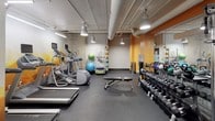 Located at 1211 Dexter Avenue North in Seattle's South Lake Union neighborhood, this fitness center offers a well-equipped, single-floor workout facility designed to support a wide range of exercise routines. The space is divided into distinct zones, each tailored to specific training needs. The cardio area features multiple treadmills set against a striking geometric mountain-themed wall mural in warm amber and gold tones, creating an energizing focal point. Nearby, elliptical trainers and additional cardio machines line the space alongside full-length mirrors that visually expand the room. A dedicated cycling area houses stationary bikes equipped with digital screens, also backed by floor-to-ceiling mirrors and vibrant orange accent walls. The free weights zone provides a comprehensive dumbbell rack alongside a flat workout bench, catering to strength training. A functional training section rounds out the facility, stocked with stability balls, resistance bands, medicine balls, and foam rollers on organized racks. Throughout the space, exposed ductwork and industrial-style pendant lighting lend an urban aesthetic, while durable rubber flooring ensures safety and practicality. The overall design balances functionality with visual energy, making it a motivating environment for all fitness levels.