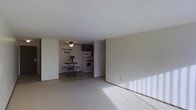 This single-floor space offers a practical and comfortable layout designed for everyday living. The spacious living area is finished with soft wall-to-wall carpeting that extends throughout, creating a warm and cohesive feel underfoot. Natural light filters into the room, casting bright patterns across the floor and lending the space an open, airy ambiance. A ceiling fan is centrally positioned to provide year-round comfort and air circulation.

Adjacent to the living area is a cozy dining space, accommodating a small bistro-style table and stools — an inviting spot for casual meals. The kitchen is efficiently designed and flows naturally from the dining area, featuring white appliances including a range and over-the-range microwave, complemented by dark upper cabinetry that adds a subtle contrast to the neutral tones of the surrounding walls. The clean, light-colored walls throughout create a bright backdrop that makes the space feel open and ready to personalize. The overall design is straightforward and functional, making it a welcoming and versatile living environment.