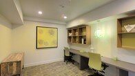 This single-floor workspace is thoughtfully designed to support productivity in a clean, modern environment. The room centers around a long, built-in desk that runs along one wall, providing ample surface area for multiple users to work side by side. Ergonomic task chairs in a muted yellow-green tone complement the neutral color palette of the space, adding a subtle pop of color without overwhelming the overall aesthetic.

Above the desk, an open-compartment wood shelving unit offers organized storage for books, décor, and office essentials, keeping the workspace tidy and functional. The crisp white walls are accented by a bold, large-format abstract artwork featuring a warm yellow background with hand-drawn floral motifs, lending the room an artistic and welcoming character.

Lighting is a highlight of the space, with recessed ceiling fixtures providing even, ambient illumination throughout. A distinctive globe pendant light in a soft green hue adds a decorative touch and visual interest. The flooring features a patterned carpet that introduces texture and warmth underfoot. A mirrored wall element reflects light and creates a greater sense of openness. Overall, the space balances practical functionality with a refined, contemporary design sensibility.