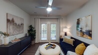 This single-floor residence offers a thoughtfully designed layout that balances comfort and modern style throughout. The kitchen is a standout feature, equipped with crisp white shaker cabinetry, dark countertops, a classic subway tile backsplash, and a full suite of stainless steel appliances including a French door refrigerator, range, microwave, and dishwasher. Hardwood-style flooring runs through the kitchen and dining area, where a compact dining set sits adjacent to the kitchen, making everyday meals convenient and inviting. The living room provides a relaxed atmosphere, highlighted by ceiling fan lighting, neutral walls, and a set of French doors that open to an outdoor area, allowing natural light and greenery views to enhance the space. The bedroom features plush carpeting, large windows framed by bold drapes, a ceiling fan, and generous room for furnishings, creating a restful retreat. A secondary room with soft carpet and neutral tones offers flexibility as an additional bedroom or versatile space. Completing the home is a dedicated laundry room with a full-size washer and dryer and overhead shelving for added storage. The overall aesthetic is clean, contemporary, and functional throughout.