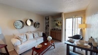 This single-floor residence centers around a well-appointed living area finished with wall-to-wall carpeting and a textured ceiling that adds subtle dimension overhead. The main lounge is anchored by a plush, neutral-toned sofa dressed with layered throw pillows, complemented by a warm wood coffee table that grounds the seating arrangement. Decorative circular mirrors line one wall, adding visual interest and depth to the space.

A standout built-in feature along the back wall combines open shelving with a lower wood-cabinet wet bar, ideal for displaying glassware and entertaining essentials. Glass shelving above the bar area provides additional display space, while the cabinetry below offers concealed storage. Natural light filters generously into the room through a sliding glass door, which opens onto an outdoor balcony, creating a seamless connection between the interior and exterior. The warm, neutral color palette throughout the living area—soft beiges and earthy wood tones—contributes to a cohesive and inviting ambiance suited for both relaxing and hosting guests.