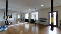This single-floor fitness space offers a clean, functional environment designed for a variety of workout styles. The room features warm hardwood flooring throughout, providing a durable and attractive surface suitable for both strength training and bodyweight exercises. A large floor-to-ceiling mirror spans one full wall, visually expanding the space and allowing users to monitor their form during workouts.

Natural light filters in through multiple white-framed windows along the exterior wall, creating a bright and inviting atmosphere complemented by recessed ceiling lighting. The space is equipped with TRX suspension training systems, suspended from the ceiling on yellow straps, offering versatile functional fitness options. A dedicated kettlebell rack holds an organized selection of kettlebells in varying weights, while stacked weight plates are neatly arranged nearby, supporting a range of strength training routines.

The neutral color palette—white walls, light trim, and natural wood tones—contributes to an open, airy ambiance. The overall layout is thoughtfully arranged to maximize open floor space for movement, making this fitness area well-suited for both individual training sessions and small group workouts.