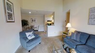 This single-floor residence offers a well-designed open-concept layout that seamlessly connects the living, dining, and kitchen areas into one cohesive and functional space. The living area is generously sized, featuring plush wall-to-wall carpeting and soaring ceilings that lend a sense of openness and airiness. Comfortable seating, including upholstered armchairs and a sofa, create an inviting atmosphere centered around a glass-top coffee table.

Adjacent to the living space is a dedicated dining area, furnished with a round glass-top table and stylish metal chairs, making it ideal for both casual meals and entertaining guests. The space is accented with warm, neutral tones on the walls and subtle decorative touches, including framed artwork and indoor greenery.

The kitchen features a convenient pass-through counter that opens to the main living and dining areas, promoting interaction and ease of use. White cabinetry, under-cabinet lighting, and ample counter space round out the functional kitchen design.

Throughout the space, recessed lighting and a combination of natural and artificial light maintain a bright and welcoming ambiance. The neutral color palette and soft carpeting tie the rooms together, creating a harmonious and comfortable living environment suitable for everyday living.