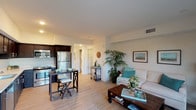 This model unit at Vista Village offers a well-designed, single-floor open-concept layout that seamlessly connects the kitchen, dining, and living areas. The kitchen is outfitted with rich dark cabinetry, light quartz-style countertops, stainless steel appliances including a refrigerator, dishwasher, range, and microwave, and a double sink. Light wood-style flooring runs throughout, creating a clean and cohesive aesthetic. The dining area accommodates a bistro-style table adjacent to the kitchen, making everyday meals convenient and casual. The living room is bright and airy, featuring large windows that invite natural light and offer views of the surrounding greenery. The space is comfortable and generously proportioned. Two bedrooms are included, each with sliding closet doors, ceiling fans, and windows overlooking outdoor landscaping. The full bathroom features a dark vanity with a marble-style countertop, a large mirror, and a shower-tub combination. A hallway closet houses a stacked in-unit washer and dryer, adding everyday convenience. Throughout the unit, neutral walls and light flooring create a calm, inviting atmosphere well-suited for modern living.