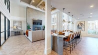 This elegantly designed single-floor communal space offers a seamless open-plan layout that integrates multiple distinct functional zones. The space is unified throughout by striking geometric parquet wood flooring, which adds warmth and visual interest underfoot.

The lounge area anchors one end of the space with a dramatic floor-to-ceiling stacked stone accent wall, serving as a backdrop for a large wall-mounted television and a sleek fireplace below. Comfortable seating arrangements, including plush sofas and accent chairs, create an inviting setting for relaxation and social gathering. Exposed wood beam detailing on the ceiling and an elegant chandelier contribute to the room's refined yet welcoming ambiance.

A long marble-topped kitchen island with leather bar stools runs through the heart of the space, illuminated by a row of industrial-style pendant lights. The adjacent full kitchen features white shaker cabinetry, dark stone countertops, a bold chevron-patterned tile backsplash, stainless steel appliances, and a built-in wine refrigerator — making it well-equipped for entertaining.

At the opposite end, floor-to-ceiling windows flood the space with abundant natural light and offer open views of the surrounding outdoor courtyard and community grounds. A casual dining area with round tables and leather chairs is positioned to take full advantage of these expansive views, creating a bright and airy atmosphere throughout.