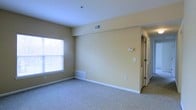 This single-floor residential space offers a clean, functional layout designed for comfortable everyday living. The main living area is finished with wall-to-wall carpeting in a neutral tone, providing a soft and inviting atmosphere throughout. Warm beige walls complement the light color palette, creating a cohesive and understated aesthetic.

Natural light enters the living area through a well-positioned window fitted with a white blind, brightening the space during the day. The ceiling is smooth and bright white, contributing to an open, airy feel. Overhead, a centrally located smoke detector and a ceiling-mounted HVAC vent ensure comfort and safety.

Climate control is well-considered, with both ceiling and floor-level HVAC vents distributed throughout the room, allowing for efficient air circulation year-round. A wall-mounted thermostat provides convenient temperature management.

Toward the rear of the living space, bifold doors reveal a closet, offering practical built-in storage. White trim and baseboards frame the walls cleanly, adding a crisp finishing detail throughout. The overall layout is straightforward and versatile, lending itself well to a variety of furniture arrangements and personal styles. The space presents a move-in ready environment with all the essential features in place.