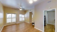 This single-floor residence offers a thoughtfully designed layout centered around a bright and open living area. The space features warm hardwood-style flooring throughout, which complements the neutral beige wall tones to create a clean, cohesive aesthetic. Multiple windows allow an abundance of natural light to fill the room, enhancing the airy and welcoming atmosphere. A ceiling fan with an integrated light fixture is centrally mounted, providing both ventilation and additional illumination. The living area flows seamlessly into adjacent rooms through a wide interior archway, suggesting a well-connected floor plan designed for ease of movement. White baseboards and trim provide a crisp contrast to the warm wall tones, adding a polished finish to the interior. The open passage visible beyond the living space hints at additional rooms, further contributing to the sense of spaciousness. The overall design aesthetic is minimal and practical, with clean lines and a neutral palette that provides a versatile backdrop suitable for a variety of furnishing styles. The combination of natural light, durable flooring, and open sightlines makes this a functional and inviting single-level living space.