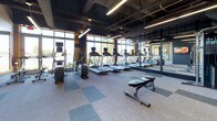 Located at 101 Avenue of the Arts in Wilmington, Delaware, this fitness center delivers a well-equipped, modern workout environment designed to meet a wide range of exercise needs. The space showcases an industrial-inspired aesthetic, with exposed ceilings revealing ductwork and structural beams painted in dark tones, complemented by sleek linear pendant lighting that illuminates the floor below. Original brick accent walls add warmth and texture, creating a striking contrast against the contemporary finishes throughout.

The cardio zone is lined with a row of treadmills and elliptical machines, each equipped with individual monitors. Floor-to-ceiling windows along one wall flood the area with natural light and offer views of the surrounding landscape, enhancing the workout experience. Full-length mirrors with bold yellow frames run the length of an adjacent wall, visually expanding the space.

The strength training area is equally well-appointed, featuring a comprehensive dumbbell rack with a wide weight range, adjustable benches, stability balls, resistance accessories, and a cable machine system. A wall-mounted television provides entertainment throughout the space. The flooring consists of modular carpet tiles in a checkered pattern of light and dark gray, providing both comfort and durability. A water fountain is conveniently accessible within the space.