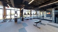Located at 101 Avenue of the Arts in Wilmington, Delaware, this fitness center delivers a well-equipped, modern workout environment designed to meet a wide range of exercise needs. The space showcases an industrial-inspired aesthetic, with exposed ceilings revealing ductwork and structural beams painted in dark tones, complemented by sleek linear pendant lighting that illuminates the floor below. Original brick accent walls add warmth and texture, creating a striking contrast against the contemporary finishes throughout.

The cardio zone is lined with a row of treadmills and elliptical machines, each equipped with individual monitors. Floor-to-ceiling windows along one wall flood the area with natural light and offer views of the surrounding landscape, enhancing the workout experience. Full-length mirrors with bold yellow frames run the length of an adjacent wall, visually expanding the space.

The strength training area is equally well-appointed, featuring a comprehensive dumbbell rack with a wide weight range, adjustable benches, stability balls, resistance accessories, and a cable machine system. A wall-mounted television provides entertainment throughout the space. The flooring consists of modular carpet tiles in a checkered pattern of light and dark gray, providing both comfort and durability. A water fountain is conveniently accessible within the space.