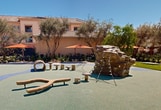 This expansive outdoor area serves as a central hub for community engagement and recreation. The space is anchored by a visually striking playground, featuring a soft, rubberized surface in soothing shades of green and blue. A unique, log-shaped play structure offers children an imaginative climbing experience. Surrounding the play area, mature olive trees provide natural shade and a Mediterranean ambiance. The landscape design incorporates a harmonious blend of drought-resistant plants and well-manicured lawns, creating a sustainable and aesthetically pleasing environment. Multiple seating options are available throughout the space, including benches and tables sheltered by vibrant orange umbrellas, encouraging social interaction and outdoor relaxation. Vintage-style lampposts add charm and illuminate the area for evening use. A welcoming signage near the entrance guides visitors, while the backdrop of warm-toned residential buildings creates a sense of enclosure and community. This thoughtfully designed space caters to various activities and age groups, promoting a healthy, active lifestyle and fostering a strong sense of community among residents.