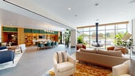 Located at 4400 Calvert Rd in College Park, Maryland, this single-floor amenity space is designed with a contemporary aesthetic that balances comfort and sophistication. The open-plan layout connects several distinct functional zones, all unified by large-format tile flooring and a bright, airy palette. Floor-to-ceiling windows flood the interior with natural light and provide direct views of an outdoor terrace, where lounge seating and a pool are visible beyond.

The main lounge area features a curated arrangement of cream swivel chairs, a neutral sofa, and a vibrant abstract area rug, anchored by a striking suspended fireplace. A statement abstract painting adorns one wall, adding an artistic focal point. Adjacent to the lounge, a generously sized dining table surrounded by teal upholstered chairs sits beneath an elegant multi-globe brass chandelier, with lush tropical plants and sliding glass doors opening directly to the terrace.

The kitchen and bar area is defined by warm wood shelving with green-tiled accents, integrated display lighting, and a flat-screen TV. A curved entryway alcove features a custom channel-tufted blue banquette, textured woven wall panels, and globe sconces. A separate nook with a rich wood accent wall houses an additional flat-screen display, completing this well-appointed communal space.