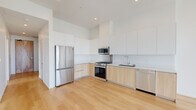 Located at 200 W Ocean Blvd in the heart of Long Beach, CA, this modern 2-bedroom, 3-bathroom residence offers a clean, contemporary design defined by an open layout, light hardwood flooring, and crisp white walls throughout. The two living areas are bathed in natural light, courtesy of expansive floor-to-ceiling windows that frame elevated views of the surrounding Long Beach cityscape, including palm tree-lined streets and the urban skyline. The kitchen is thoughtfully appointed with two-tone cabinetry — white upper cabinets paired with warm wood lower cabinets — complemented by a suite of stainless steel appliances including a gas range, built-in microwave, dishwasher, and French door refrigerator, all set against light wood flooring. The primary bedroom offers generous proportions, a built-in closet, and direct access to one of the three bathrooms, which features large-format grey tile, a glass-enclosed walk-in shower with a handheld fixture, and a lighted mirror vanity with wood cabinetry. The second bedroom is bright and airy with a tall window overlooking the city. A hallway connects all rooms efficiently, with a dedicated closet for added storage. The overall ambiance is modern, light-filled, and urban.