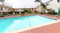 This property's outdoor common area centers around a generously sized swimming pool set within a courtyard-style layout, enclosed by a dark metal safety fence. The pool deck is finished in a deep terracotta-toned concrete surface, providing a warm, inviting contrast to the clear blue water of the pool. Lounge chairs with patterned cushions and wrought iron frames are positioned poolside, offering comfortable spaces for relaxation. A large shade umbrella extends over a round wrought iron table and matching chairs, creating a shaded outdoor dining and gathering area. Decorative potted palms and flowering plants add tropical character throughout the space. The surrounding multi-story building features exterior walkways, stairwells, and balconies that overlook the courtyard. Well-maintained green lawns border the pool area, complemented by neatly trimmed hedges and mature palm trees that provide a lush, landscaped backdrop. The overall ambiance is that of a resort-inspired communal outdoor space, designed for leisure and social activity within a residential complex setting.