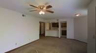 This single-story space features an open-concept layout that seamlessly connects the living area to the kitchen. The spacious living room is finished with wall-to-wall carpeting and neutral white walls, creating a clean and versatile backdrop. A ceiling fan with an integrated light fixture is centrally mounted, providing both air circulation and ambient lighting to the room.

The kitchen is partially open to the living area and is defined by a raised breakfast bar that serves as a natural dividing element. Medium-toned wood cabinetry lines the kitchen walls, offering ample storage, while dark countertops provide contrast. Recessed lighting illuminates the kitchen workspace. A bank of lower cabinetry is visible to the right side of the kitchen, suggesting additional prep or storage space.

A hallway visible from the living area leads to additional rooms within the unit. HVAC vents are positioned throughout the ceiling, indicating a central air and heating system. The overall ambiance is straightforward and functional, with a neutral color palette that lends itself well to a variety of furnishing styles.