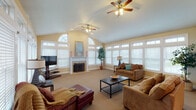 This single-floor space is thoughtfully designed with distinct living, dining, and kitchen areas that flow seamlessly together. The expansive living room is a standout feature, boasting vaulted ceilings accented by two ceiling fans and an abundance of windows fitted with white blinds, allowing generous natural light to fill the room. A traditional fireplace with a white mantle serves as a focal point, flanked by an arched transom window. Plush carpeting and ample seating space make the area ideal for relaxation or entertaining.

The kitchen is well-appointed with warm honey-toned cabinetry, stone-look countertops, and a full suite of stainless steel appliances, including a range, over-the-range microwave, and dishwasher. Light ceramic tile flooring extends from the kitchen into the dining area, which is brightened by large windows with transom details on two walls. A chandelier overhead and a round wooden dining table with wrought iron chairs complete the inviting dining space. A breakfast bar connects the kitchen to the dining area, offering additional casual seating. Together, these areas create a cohesive and functional living environment.