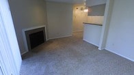 This single-floor residential space offers a practical and comfortable layout designed for everyday living. The main living area is finished with wall-to-wall carpet, creating a warm and quiet underfoot experience throughout the space. A classic fireplace with a tiled surround serves as a natural focal point along one wall, adding character and a cozy ambiance to the room. Neutral-toned walls throughout contribute to a clean, versatile aesthetic that adapts easily to a variety of décor styles. A ceiling fan is centrally positioned overhead, providing both light and air circulation to the living area. The space flows seamlessly toward a kitchen area, which is defined by a raised pass-through counter with a dark laminate top — a functional architectural detail that connects the kitchen to the main living space while maintaining a sense of separation. This open-concept approach enhances the overall sense of space and encourages an easy, social flow between areas. The layout is straightforward and efficient, making it well-suited for those seeking a low-maintenance, single-level living environment with classic residential features.
