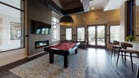 Located at 161 Grande Oaks Dr in Charleston, SC, this single-floor community clubhouse delivers an upscale, lodge-inspired atmosphere designed for socializing and recreation. The space is anchored by rich dark hardwood-style tile flooring that flows seamlessly throughout, complemented by bold dark trim accents and warm neutral wall tones that create a cohesive, sophisticated aesthetic.

The game and entertainment area is a standout feature, housing a red felt billiards table beneath a striking dome pendant light, a foosball table, and a wall-mounted flat-screen TV above a sleek linear electric fireplace. Plaid drapery and large windows flood the space with natural light while framing views of the surrounding greenery.

A dedicated card and poker area features a round gaming table surrounded by rich burgundy leather chairs, set beneath coffered ceilings with dark-stained beam detailing and another bold dome pendant fixture. Nearby, a more casual seating area with a rectangular table and upholstered chairs provides space for meetings or informal gatherings.

A sliding barn door separates zones within the open layout, adding a charming architectural detail. The space also includes lounge seating with teal accent chairs, a media room glimpsed through interior windows, and direct access to an outdoor area via French doors, enhancing the sense of openness throughout.