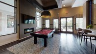 Located at 161 Grande Oaks Dr in Charleston, SC, this single-floor community clubhouse delivers an upscale, lodge-inspired atmosphere designed for socializing and recreation. The space is anchored by rich dark hardwood-style tile flooring that flows seamlessly throughout, complemented by bold dark trim accents and warm neutral wall tones that create a cohesive, sophisticated aesthetic.

The game and entertainment area is a standout feature, housing a red felt billiards table beneath a striking dome pendant light, a foosball table, and a wall-mounted flat-screen TV above a sleek linear electric fireplace. Plaid drapery and large windows flood the space with natural light while framing views of the surrounding greenery.

A dedicated card and poker area features a round gaming table surrounded by rich burgundy leather chairs, set beneath coffered ceilings with dark-stained beam detailing and another bold dome pendant fixture. Nearby, a more casual seating area with a rectangular table and upholstered chairs provides space for meetings or informal gatherings.

A sliding barn door separates zones within the open layout, adding a charming architectural detail. The space also includes lounge seating with teal accent chairs, a media room glimpsed through interior windows, and direct access to an outdoor area via French doors, enhancing the sense of openness throughout.