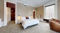 Located at 888 Delaware Ave in Buffalo, NY, this thoughtfully appointed one-bedroom, one-bathroom unit blends historic charm with modern comfort. The spacious bedroom is the centerpiece of the layout, offering generous proportions with warm taupe walls, ornate crown molding, and tall ceilings that lend an air of elegance. A striking white marble decorative fireplace anchors one wall, complemented by a classic white radiator nearby. Large double-hung windows flood the room with natural light and provide views of the surrounding neighborhood. A ductless mini-split system ensures year-round climate control. The bathroom continues the refined aesthetic with marble-topped dual vanity cabinetry, large-format tile flooring, a glass-enclosed shower, and a bright window overlooking the streetscape. A dedicated laundry and kitchenette area provides added convenience with a compact sink, appliances, and storage. The hallway features elegant wainscoting and brass wall sconces, connecting the rooms with architectural continuity. Rich dark wood doors throughout add warmth and historic character to this meticulously detailed space.