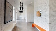 This single-floor space presents a contemporary lobby designed with a clean, urban aesthetic that balances industrial character with modern refinement. The defining architectural feature is the white-painted brick that lines every wall, lending a bright yet textured backdrop throughout the space. Polished concrete floors reflect natural light and reinforce the industrial sensibility, while contributing to an open, airy atmosphere.

Generous windows along one side flood the interior with natural light and offer views of lush greenery outside, creating a welcoming sense of connectivity to the outdoors. A bold orange bench serves as a striking focal point, introducing a vibrant pop of color against the all-white palette, while a sleek white built-in bench provides additional seating on the opposite side.

A dramatic cluster of black industrial pendant lights hangs from the ceiling, serving as both a functional and sculptural centerpiece. The high ceilings amplify the sense of volume and openness throughout the corridor-style layout.

An entire wall is dedicated to a large array of built-in white mailboxes, seamlessly integrated into the brick surround for a cohesive look. Carefully curated framed artwork adorns the walls, adding personality and visual interest. A small white service counter near the entrance completes the functional amenities of this well-appointed communal lobby space.