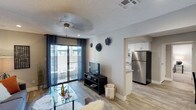 This single-floor residence offers a well-designed open-concept layout that seamlessly connects the living area to the kitchen. The living room is bright and welcoming, featuring light wood-style flooring, a ceiling fan, and a textured ceiling. A sliding glass door floods the space with natural light and leads to a private balcony, perfect for outdoor relaxation. Decorative wall accents add a stylish, personalized touch to the neutral-toned walls, complemented by navy blue curtains that frame the balcony entrance. The kitchen is visible from the living area and showcases white cabinetry, a stainless steel refrigerator, a light-toned countertop, and clean, modern finishes throughout. The open pass-through design between the kitchen and living space enhances the sense of flow and spaciousness. Overall, the residence strikes a balance between functional design and contemporary style, making it a comfortable and practical living environment with a cohesive aesthetic carried throughout its shared spaces.