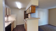 This single-story unit offers a functional and straightforward layout designed for comfortable everyday living. The kitchen is configured as a galley, featuring light wood cabinetry on both sides, a white electric range, a black dishwasher, a refrigerator, and a stainless steel sink. A peninsula with a laminate countertop extends outward, creating a breakfast bar that connects the kitchen to the adjacent living area. Overhead cabinetry with an integrated range hood provides additional storage above the cooking surface. Flush-mounted ceiling fixtures illuminate the kitchen, which is finished with dark hardwood-style flooring throughout.

The living area is carpeted and receives natural light through a sliding glass door fitted with vertical blinds. A pendant light fixture adds overhead illumination, and a louvered closet houses a stacked washer and dryer unit. The bedrooms are carpeted as well, each featuring windows with blinds and louvered closet doors providing accessible storage. Textured ceilings are consistent throughout the unit.

Both bathrooms share a similar design, each equipped with a single-sink vanity with wood cabinetry, a wall mirror with a multi-bulb light bar, a toilet, and a shower-tub combination with a curtain rod and tile surround. Dark hardwood-style flooring carries through the bathrooms, complementing the overall neutral color palette of the unit.