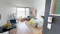 This thoughtfully designed single-floor unit showcases a contemporary open-concept layout where the living, dining, and sleeping areas flow seamlessly together. Rich hardwood-style flooring runs throughout, creating a warm and cohesive aesthetic. The living area benefits from expansive floor-to-ceiling corner windows that flood the space with natural light and offer open views of the surrounding urban streetscape, making it an ideal spot for relaxing or working.

The kitchen is efficiently laid out with dark lower cabinetry contrasted by crisp white upper cabinets and open shelving. It is equipped with stainless steel appliances including a range, microwave, dishwasher, and refrigerator, complemented by a subway tile backsplash and a deep stainless steel sink with a modern pull-down faucet. A compact dining nook adjacent to the kitchen provides a functional space for meals.

The sleeping area is generously proportioned, accommodating a full-sized bed with room to spare, and features additional windows that continue the theme of abundant natural light. A dedicated workspace with a desk and chair adds practical functionality.

The bathroom features warm hardwood-style flooring, a dark vanity with ample counter space, overhead cabinetry for storage, and clean white walls that keep the space feeling bright and airy.