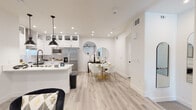 This well-appointed single-floor residence showcases a cohesive modern design throughout, anchored by light wood-look flooring, crisp white walls, and recessed lighting that create a bright and airy atmosphere. The open-concept kitchen is outfitted with white shaker cabinetry, glass-front upper cabinets, a subway tile backsplash, a stainless steel wall-mounted range hood, and a full suite of stainless steel appliances including a large side-by-side refrigerator and slide-in range. A central island provides additional prep and seating space, while a sleek pendant light adds a contemporary accent. The kitchen flows seamlessly into the dining area, where a round marble-top table and modern white chairs are complemented by a pair of round decorative mirrors and a dark accent console. The living room offers generous space, complete with a ceiling fan, wall-mounted TV, and access to a bright window frontage framed by neutral drapes. The bedrooms feature plush carpeting, ceiling fans, ample natural light, wall-mounted TVs, and walk-in closet access. Bathrooms are finished with light tile flooring, shiplap-style doors, and clean white vanities with matte black hardware. A dedicated laundry room with a full-size washer and dryer rounds out this thoughtfully designed residence.