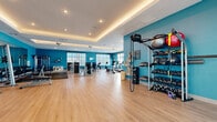 Located at 2500 Conserve Cir in Apopka, Florida, this expansive fitness center offers a well-appointed, single-floor layout designed to support a wide range of workout routines. The space is defined by its bold teal accent walls, light hardwood-style flooring, and a striking tray ceiling with warm cove lighting that adds a polished, modern feel throughout.

Large windows line multiple walls, flooding the interior with abundant natural light and offering views of the surrounding Florida landscape, including palm trees and the outdoor amenity areas. Ceiling fans and recessed lighting ensure comfort and visibility across the entire floor.

The fitness center is comprehensively equipped with dedicated zones for various training styles. A cardio section features treadmills, ellipticals, stationary bikes, and a stair climber. A strength training area includes a full rack of dumbbells backed by a large wall mirror, a functional trainer, cable machines, and a multi-use power rack. A separate functional fitness zone provides TRX suspension trainers, battle ropes, kettlebells, medicine balls, and BOSU trainers. Selectorized weight machines round out the strength offerings.

A wall-mounted television, a water station, and open shelving with accessories contribute to the overall usability and comfort of the space. Glass doors open to an adjacent outdoor area, enhancing the airy, connected atmosphere of this well-designed community fitness facility.
