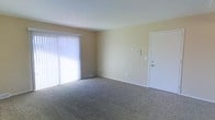 This single-floor space features a straightforward and functional layout designed for everyday living. The main living area is finished with wall-to-wall carpet in a neutral tan tone, providing a comfortable underfoot feel throughout the room. Walls are painted in a soft, warm beige, contributing to a clean and understated aesthetic.

A prominent feature of the living area is the large sliding glass door dressed with vertical blinds, which allows an abundance of natural light to fill the room. A floor-level heating vent is positioned near the sliding door, ensuring efficient climate control. The room also includes a standard interior door along the adjacent wall, providing access to additional areas of the space. Wall-mounted utility panels—likely a thermostat and light switch—are neatly placed nearby.

The ceilings are a standard height, keeping the space feeling practical and easy to furnish. Overall, the space presents a neutral, move-in-ready environment with simple finishes and ample natural light, making it well-suited for a variety of furnishing styles and uses.