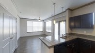 This single-story residence showcases an open-concept layout that connects the kitchen and living areas into one cohesive, functional space. The kitchen is equipped with dark espresso cabinetry, granite countertops with subtle speckled detailing, and a kitchen island that doubles as a breakfast bar. A stainless steel sink is positioned on the island, complemented by two elegant pendant lights that hang above, providing focused task lighting. Dark appliances, including a dishwasher, coordinate with the cabinetry for a unified aesthetic. Crown molding along the upper walls adds a refined architectural detail throughout the space. The flooring features smooth, wood-look planks in a neutral tone that extend seamlessly from the kitchen into the living area, enhancing the sense of continuity. The living area is generously proportioned, bathed in natural light from multiple large windows dressed with horizontal blinds. A ceiling fan is centrally positioned in the living area, offering both lighting and air circulation. The overall ambiance is clean, contemporary, and bright, with a neutral color palette that lends itself to a variety of interior styles.