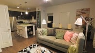 This single-floor space showcases an open-concept design that seamlessly connects the living area and kitchen, creating an inviting and functional layout. The living area is anchored by a tufted sofa in a neutral tone, accented with vibrant green and coral throw pillows, adding pops of color to the warm, earthy palette. A stylish floor lamp provides soft ambient lighting, complementing the recessed ceiling lights. A low decorative ledge along one wall displays tasteful accents, adding depth to the space.

The kitchen features dark espresso cabinetry that contrasts elegantly with light granite countertops on the peninsula, which doubles as a breakfast bar with bar stool seating. Stainless steel appliances, including a microwave and range, add a modern touch. Pendant lighting hangs above the peninsula, enhancing both functionality and style.

Rich hardwood-style flooring flows throughout, grounding the space with warmth and continuity. A bold black-and-white patterned area rug defines the living zone. A doorway visible from the living area hints at additional rooms beyond. The overall ambiance is cozy yet contemporary, well-suited for both relaxing and entertaining.