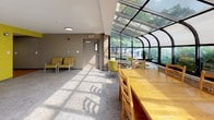 Located on Page Terrace in Stoughton, Massachusetts, this single-story space is thoughtfully designed to accommodate both relaxation and functional gathering. The interior is characterized by a warm, contemporary palette featuring taupe and bold yellow accent walls, carpeted flooring in the lounge areas, and hardwood flooring near the kitchenette.

The main lounge area is furnished with tan leather armchairs and a gray upholstered sofa, arranged to encourage conversation and comfort. Natural light streams in through windows fitted with vertical blinds, casting patterned light across the carpet. A separate seating nook provides additional intimate gathering space with matching yellow-green chairs and a dining table.

A glass-enclosed sunroom is a standout architectural feature, with a curved glazed roof and full-length windows that frame views of the surrounding greenery. Exposed brick detailing along the interior walls adds texture and warmth to this bright space, which is furnished with a long wooden dining table and coordinating chairs.

The compact kitchenette is well-appointed with white cabinetry, laminate countertops, a stainless steel sink, full-size refrigerator, microwave, and a single-serve coffee maker, providing convenient amenity support for occupants.