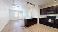 This single-floor residence offers a thoughtfully designed open-concept layout that seamlessly connects the kitchen and living area. The kitchen is a standout feature, showcasing rich dark espresso cabinetry with sleek hardware, white countertops, and a full suite of black appliances including a refrigerator, electric range, over-the-range microwave, and dishwasher. A central kitchen island with a built-in sink and breakfast bar overhang provides both additional workspace and casual seating. Track lighting above the island adds focused illumination, complementing the warm, light-toned wood-style flooring that extends throughout the main living spaces. The living area is bright and open, benefiting from natural light through slatted blinds and a ceiling fan overhead. A private exterior door in the living area provides convenient outdoor access. The bedrooms are carpeted and well-lit, each featuring windows with blinds and ceiling fans for comfort. The bathroom is clean and functional, featuring a combined tub and shower, a single-sink vanity with dark cabinetry matching the kitchen aesthetic, and tile-style flooring. A generously sized walk-in closet with multi-tiered wire shelving provides ample storage. Overall, the space presents a cohesive, modern design with neutral tones throughout.