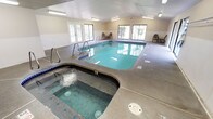 Located in Port Orchard, Washington, this single-story indoor aquatic facility offers a well-appointed space designed for year-round swimming and relaxation. The centerpiece of the facility is a generously sized indoor swimming pool, tiled in classic blue along the waterline and equipped with stainless steel entry railings on multiple sides for easy access. The pool's clear turquoise water is complemented by a speckled, slip-resistant concrete deck that surrounds it on all sides, providing ample space for poolside seating. An adjacent hot tub offers a dedicated space for hydrotherapy and relaxation, seamlessly integrated into the pool deck area. The facility is bathed in natural light through an abundance of large windows and sliding glass doors positioned along multiple walls, offering views of the surrounding outdoor greenery. Overhead, fluorescent lighting ensures the space remains well-lit at all times. Seating areas with chairs are thoughtfully placed along the perimeter for guests to rest and observe. A private rinse shower station, finished with clean white subway tile, is conveniently located near the entry point of the facility. The overall ambiance is clean, functional, and inviting, making it a practical and comfortable aquatic amenity.