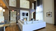 This single-floor space makes a striking architectural statement with its dramatic double-height ceilings, creating an expansive, airy atmosphere that immediately commands attention. The main lounge area is anchored by a sleek, wall-mounted fireplace topped with a flat-screen television, framed within a clean white surround that contrasts beautifully against the rich, deep brown accent walls. Large-scale contemporary artwork is displayed prominently on the upper walls, adding a gallery-like sophistication to the space.

A generously sized white sectional sofa defines the seating area, offering a refined yet inviting focal point centered around the fireplace. Floor-to-ceiling windows dressed in sheer white drapery flood the interior with natural light while providing softened views of the outdoors. Warm hardwood flooring runs throughout, grounding the space with natural texture and complementing the bold wall tones.

Thoughtfully placed wall sconces provide ambient lighting, while the open sightlines through arched doorways suggest additional rooms and functional areas beyond. The overall design aesthetic is modern and polished, balancing clean architectural lines with carefully curated décor elements.