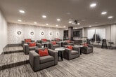 Located in Overland Park, Kansas, this single-floor screening and lounge space is thoughtfully designed for both entertainment and social gatherings. The room's centerpiece is a large projection screen framed by rich dark wood paneling, paired with a ceiling-mounted projector that delivers a true cinematic experience. Rows of generously sized, charcoal-upholstered lounge chairs are arranged throughout the space, each accented with bold red throw pillows that inject vibrant contrast against the neutral palette. Sleek round side tables with wire-frame bases are positioned between seating clusters, offering convenient surfaces for drinks and personal items.

A textured accent wall, lined with circular wall sconces, adds architectural depth and warmth along one side of the room. Recessed ceiling lighting provides even, ambient illumination throughout. Toward the rear, a wet bar area features a white countertop, dark cabinetry, under-counter refrigeration, and dramatic dark tile backsplash. High-top tables with industrial-style bar stools occupy a corner nearby, accompanied by bold black-and-white artwork. Striped, multi-toned carpeting unifies the entire floor. The overall ambiance is polished, modern, and well-suited for private screenings or community events.