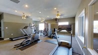 This dedicated fitness space offers a comprehensive workout environment designed to accommodate a wide range of exercise needs. The room is outfitted with multiple cardio machines, including a treadmill and elliptical trainers, as well as a multi-station strength training machine and additional weight equipment, providing users with a full-body workout experience. Neutral, earthy-toned walls complement the carpeted flooring, while protective rubber mats are placed beneath the equipment for added safety and durability. Large wall-mounted mirrors span one side of the room, enhancing the sense of openness and allowing users to monitor their form during workouts. Natural light filters in through multiple windows, creating a bright and energizing atmosphere. Overhead, decorative ceiling fans with light fixtures provide both ventilation and ambient lighting, supplemented by recessed ceiling lights that ensure the space is well-lit at all times. A wall-mounted monitor or display unit adds a functional touch for entertainment or instructional use during exercise sessions. The textured ceiling and clean, practical layout round out this well-appointed fitness facility, making it a convenient and inviting space for daily workouts.