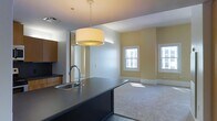 This single-floor space is designed with an open-concept layout that connects the kitchen and living area in a cohesive, functional arrangement. The kitchen features a dark-toned island with an integrated stainless steel sink and a curved gooseneck faucet, providing a striking contrast against the lighter surroundings. Warm wood cabinetry lines the kitchen wall, adding a natural element to the contemporary design. A large drum pendant light hangs above the island, serving as a central design focal point while providing ample illumination over the workspace.

The living area transitions seamlessly from the kitchen, finished with neutral wall-to-wall carpeting that adds warmth and comfort underfoot. Pale yellow walls lend a soft, inviting tone throughout the space. Two white-trimmed windows allow natural light to filter in, brightening the room and creating an airy atmosphere. The clean lines of the white baseboards and trim work complement the overall modern aesthetic.

The layout is practical and well-proportioned, making efficient use of the available space while maintaining an open, uncluttered feel. A mirrored or reflective panel near the kitchen entrance enhances the sense of depth and light within the space.
