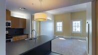 This single-floor space is designed with an open-concept layout that connects the kitchen and living area in a cohesive, functional arrangement. The kitchen features a dark-toned island with an integrated stainless steel sink and a curved gooseneck faucet, providing a striking contrast against the lighter surroundings. Warm wood cabinetry lines the kitchen wall, adding a natural element to the contemporary design. A large drum pendant light hangs above the island, serving as a central design focal point while providing ample illumination over the workspace.

The living area transitions seamlessly from the kitchen, finished with neutral wall-to-wall carpeting that adds warmth and comfort underfoot. Pale yellow walls lend a soft, inviting tone throughout the space. Two white-trimmed windows allow natural light to filter in, brightening the room and creating an airy atmosphere. The clean lines of the white baseboards and trim work complement the overall modern aesthetic.

The layout is practical and well-proportioned, making efficient use of the available space while maintaining an open, uncluttered feel. A mirrored or reflective panel near the kitchen entrance enhances the sense of depth and light within the space.