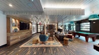 This thoughtfully designed single-floor communal lounge blends rustic and contemporary elements to create a dynamic, multi-use social space. Dark hardwood-style tile flooring runs throughout, anchoring the warm, inviting aesthetic. The white shiplap ceiling adds a contrasting brightness, while exposed brick accent walls lend an industrial, urban character to the space.

The lounge is organized into several distinct zones. A central seating area features an eclectic mix of upholstered chairs, a tufted sofa, and a patterned area rug, centered around a flat-screen television mounted within a wood-paneled media wall. Overhead, a cluster of globe pendant lights adds architectural interest.

A games area includes a full-size pool table beneath striking teal fringe pendant fixtures, as well as a shuffleboard table positioned along a brick wall. Adjacent to this, a grand piano anchors a more refined corner, complemented by framed artwork, pendant lighting, and café-style seating at marble-topped tables.

Large windows and glass doors admit generous natural light and provide views of an outdoor courtyard. A built-in bench with a marble surface and an arched window highlight create an intimate nook. Wall-mounted sconces and floor lamps complete the layered lighting throughout.
