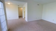 This single-floor residential space offers a straightforward and functional layout designed for comfortable everyday living. The main living area features wall-to-wall carpeting in a neutral tone, providing a warm and understated foundation throughout the space. Crisp white walls keep the interior feeling clean and open, offering a blank canvas ready for personalization.

Overhead, a textured ceiling adds a subtle architectural detail, while HVAC vents positioned throughout ensure consistent climate control across the space. A dividing wall creates a natural separation between the living area and a connecting hallway, which leads toward additional rooms including what appears to be a kitchen, visible in the background with light cabinetry and tiled or hard flooring.

The hallway area features its own overhead lighting, illuminating the transition between spaces. A baseboard heating unit is also visible along the hallway wall, indicating a supplemental heating source in addition to the central ventilation system.

Overall, the space presents a simple, well-maintained interior with a neutral color palette, practical finishes, and a cohesive single-level layout that connects the living, kitchen, and ancillary areas in an efficient and accessible manner.