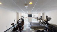 This fitness center at 195 E McMillan St in Cincinnati, Ohio, offers a well-equipped, single-floor workout facility designed to support a comprehensive cardio routine. The main exercise area is outfitted with a lineup of Landice treadmills, elliptical trainers, a stair climber, and air resistance bikes, providing a variety of options for cardiovascular training. A wall-mounted flat-screen television is positioned at the front of the room, offering entertainment during workouts. The space features a drop-panel ceiling with recessed lighting and is finished with colorful multi-toned carpet tiles in shades of green, blue, tan, and gray, giving the room a modern and clean aesthetic. Large windows and a glass sliding door flood the space with natural light and provide direct views of an adjacent outdoor pool and terrace area. A separate alcove area serves as a convenience station, equipped with a wall-mounted water bottle refill and drinking fountain as well as a shelving unit stocked with informational materials. The overall ambiance of the fitness center is functional and inviting, offering a bright, well-maintained environment suitable for daily exercise.