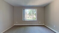This two-bedroom, one-bathroom unit is laid out across a single floor, offering a functional and comfortable living arrangement. The living room is a welcoming space featuring plush carpeting, a large picture window that frames views of mature trees and landscaped greenery, and baseboard heating along the perimeter. An adjacent area with hardwood-style flooring connects seamlessly to the rest of the unit, with a ceiling fan adding airflow and a practical touch.

The kitchen is equipped with white cabinetry, dark speckled countertops, a stainless steel sink, and a small window that brings in natural light. Storage is well-considered throughout, with built-in shelving and closet space visible in the bedroom areas.

Both bedrooms are carpeted and include windows—one with views of neighboring brick architecture and a wall-mounted air conditioning unit for climate control, and baseboard heating in each room. The bathroom is finished with mosaic tile flooring, a dark granite-style vanity countertop, a mirrored medicine cabinet, a glass-enclosed shower, and a window for ventilation and natural light. Overall, the unit provides a clean, neutral palette suited for comfortable everyday living.