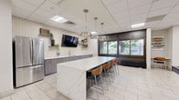 The Community Room at 115 Second Ave S in Minneapolis offers a well-appointed, open-plan gathering space designed to serve a variety of social and recreational needs. The interior is finished with large-format light tile flooring throughout, a drop ceiling with recessed panel lighting, and a neutral color palette accented by bold pops of color in the furnishings and artwork.

The kitchenette area is a standout feature, centered around a generous island with a striking white marble-style countertop and bar-height seating for several guests. Dark wood-grain cabinetry, subway tile backsplash, a stainless steel refrigerator, and a wall-mounted television complete this functional and stylish space.

The lounge area provides comfortable seating options including vibrant orange accent chairs with ottomans and modular gray sofas arranged around a coffee table, anchored by a patterned area rug. A wall-mounted television and colorful landscape artwork add visual interest to the relaxed seating zones.

A glass-walled fitness room is accessible from the main corridor, stocked with exercise equipment and fitness accessories, adding wellness functionality to the space. Large windows along one wall invite natural light and provide views to an outdoor area. The overall ambiance is modern, clean, and inviting — well-suited for community gatherings, social events, or casual everyday use.